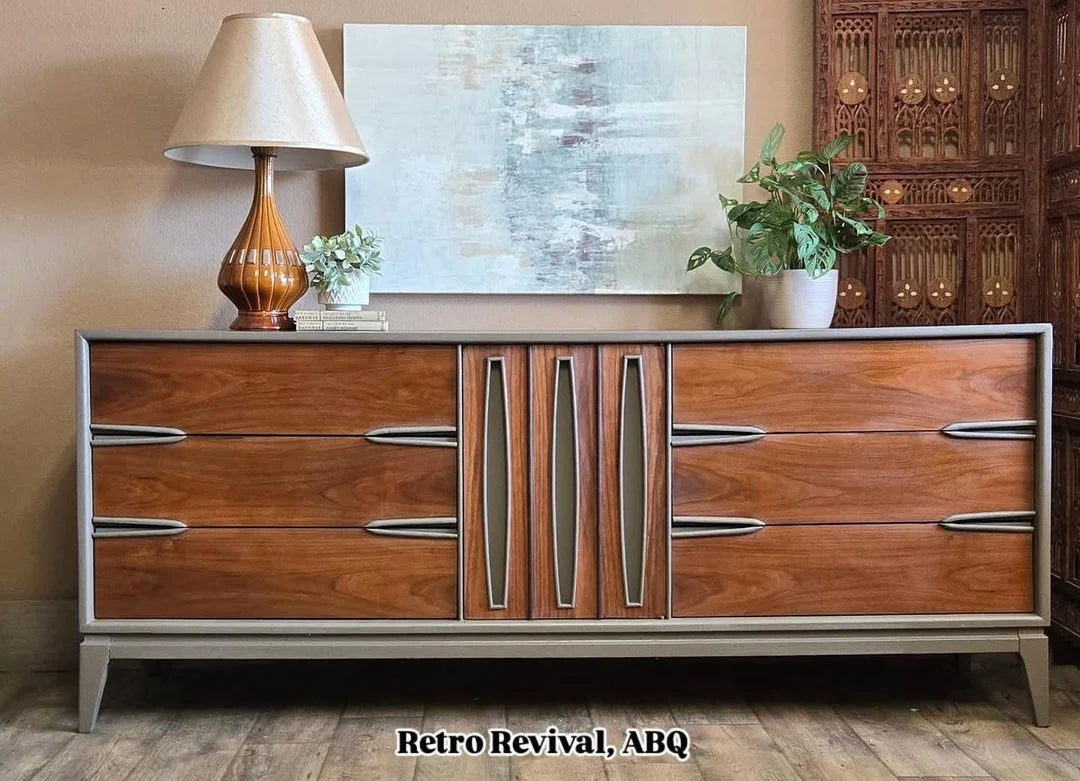 A mid-century modern wooden credenza or sideboard with six drawers, featuring warm brown wood grain and distinctive silver metallic handles, styled in a room setting with a lamp, painting, and plant.