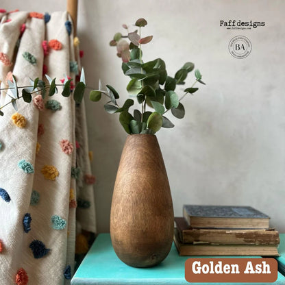 A golden ash wooden vase with a teardrop shape, filled with green eucalyptus branches, displayed on a turquoise table next to a stack of books and a patterned textile, with 'Faff designs' branding in the top right.