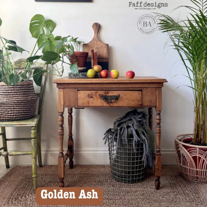 A vintage-style wooden writing desk in Golden Ash wood, styled in a cozy room setting with potted plants, a chair, and a basket, featuring a text overlay 'Golden Ash' at the bottom.
