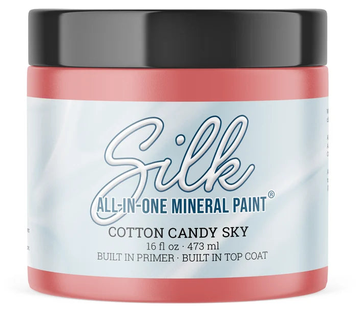 A jar of Silk All-In-One Mineral Paint in the color Cotton Candy Sky, featuring a light blue and white label with blue text, a coral pink base, and a black lid.