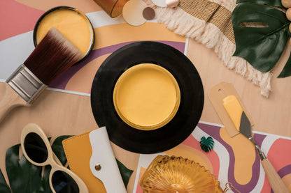 A styled flat-lay featuring a round black tray or plate filled with bright yellow cream, surrounded by decorative props including a paintbrush, sunglasses, a small fan, a notebook, and tropical leaves on a pastel abstract background.
