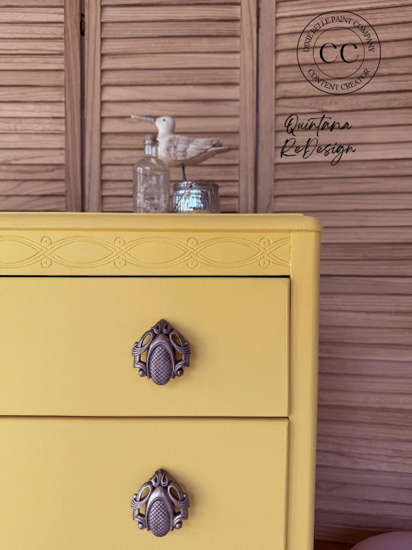 A close-up of a yellow painted dresser with ornate dark metal drawer pulls, set against a brown wooden slatted background. A seagull figurine and glass bottle sit on top.