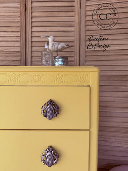 A close-up of a yellow painted dresser with ornate dark metal drawer pulls, set against a brown wooden slatted background. A seagull figurine and glass bottle sit on top.