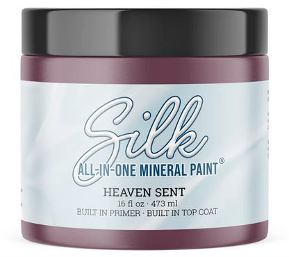 A jar of Silk All-In-One Mineral Paint in the scent Heaven Sent, featuring a dark plum purple body, a black lid, and a white label with blue text.