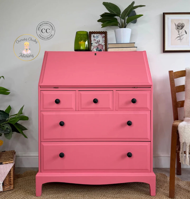 A bright pink wooden secretaire dresser with five drawers and black knobs, styled in a cozy room setting with plants, books, and a wooden chair.