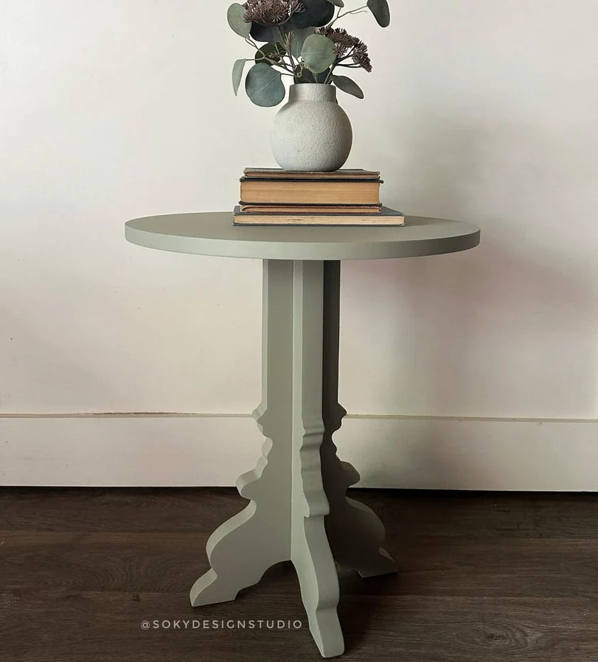 A round side table with a distressed olive green finish, styled with a white ceramic vase of eucalyptus and books on top, set against a neutral cream wall and dark wood floor.