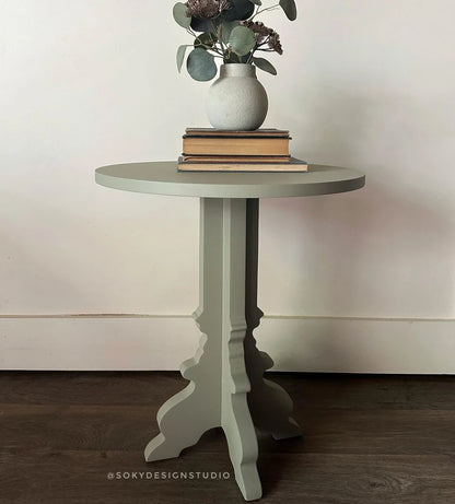 A round side table with a distressed olive green finish, styled with a white ceramic vase of eucalyptus and books on top, set against a neutral cream wall and dark wood floor.