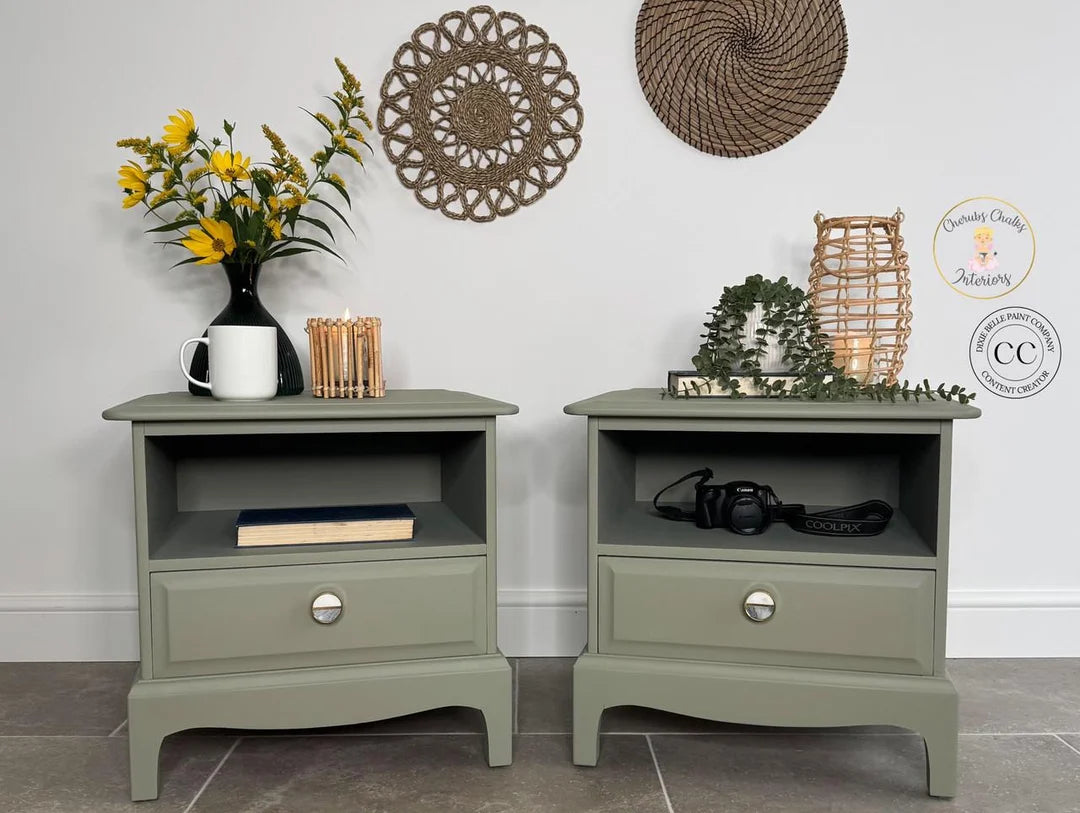 Two olive green bedside tables shown in a styled room setting with home decor, including a vase of yellow flowers, woven wall art, and decorative baskets. A circular logo for 'Carmen Clarke Furniture' is visible on the right side.