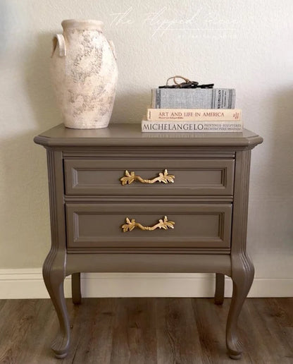 A painted nightstand with two drawers and ornate gold handles, styled with a large textured vase and books on top, set against a neutral wall and wood floor.