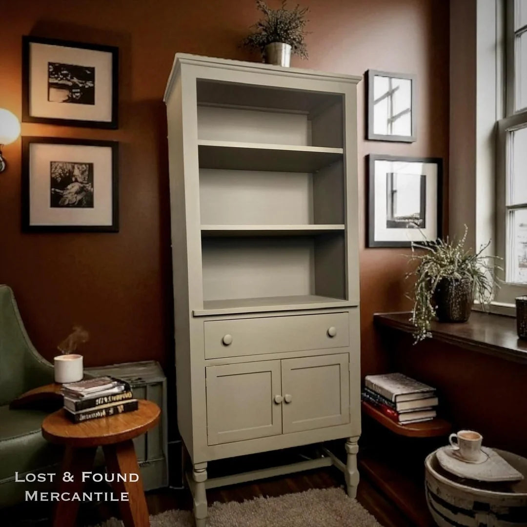 A tall, truffle coloured hutch with three open shelves and two cabinet doors with round knobs, positioned in a styled room with brown walls, framed art, a side table, and a window.
