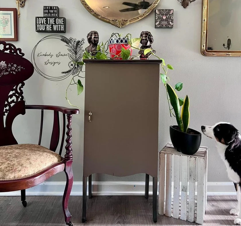 A tall, narrow cabinet in a muted taupe-brown finish stands in a styled room corner, flanked by a dark wood chair on the left and a white slatted side table with a potted plant on the right. A black-and-white dog is visible at the lower right edge.