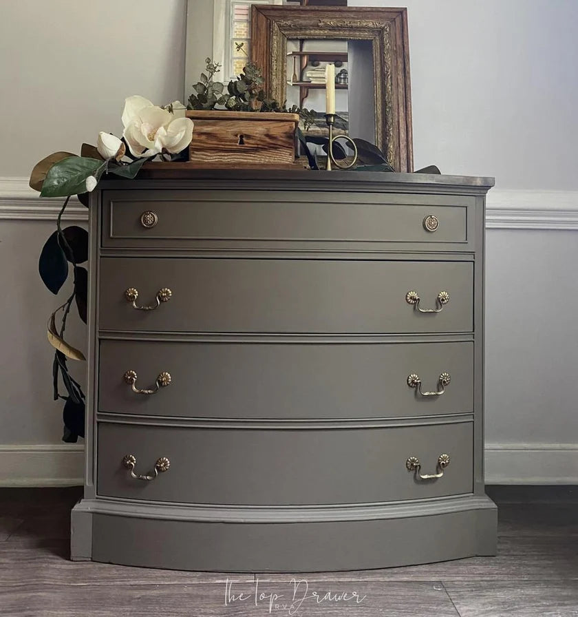 A gray three-drawer chest of drawers with brass handles, styled in a home setting with a mirror, wooden box, and flowers on top.