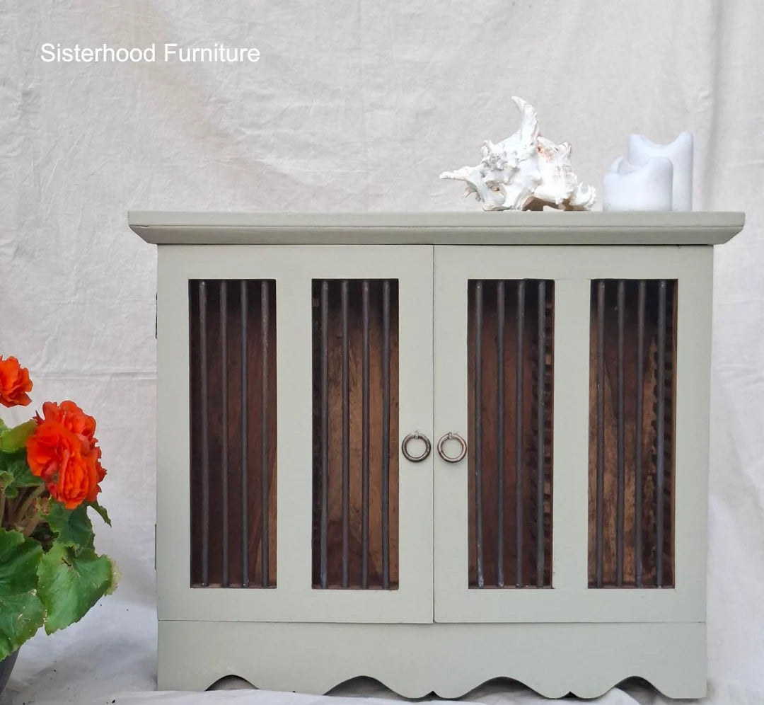 A small cabinet with a light sage green painted frame and dark brown vertical wood slat doors, shown against a white fabric backdrop. Decorative objects sit on top, and red flowers appear at the lower left.