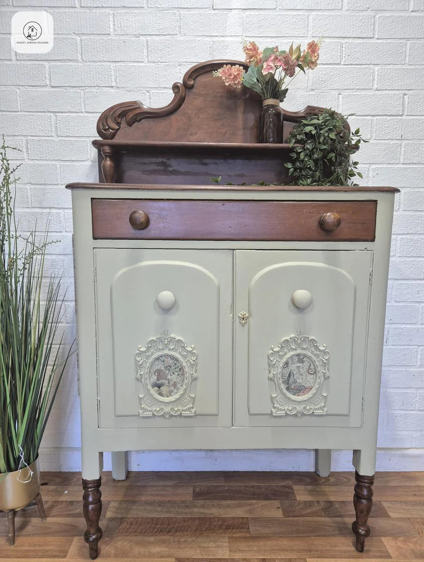 A vintage-style wooden cabinet with a distressed sage green painted lower cabinet, a dark brown drawer, and dark turned legs, styled with flowers and a plant on top against a white brick wall.